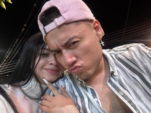 LucyAndJake with long black hair, white nails, pink top, next to Jake with a pink cap, striped shirt, and a cross earring, making a serious face, flashing a peace sign.
