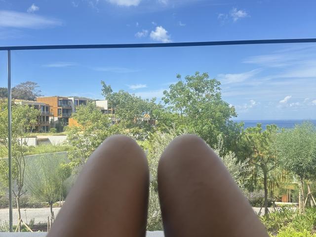 Stefanni's brown thighs are in the foreground, with her knees together. She's looking down at a clear blue sky, green trees, and a distant orange building from a balcony.