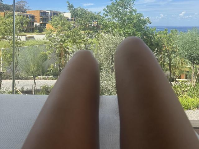 Stefanni's brown toes are in the foreground, pointing towards a green garden and blue ocean in the background, under a clear sky.