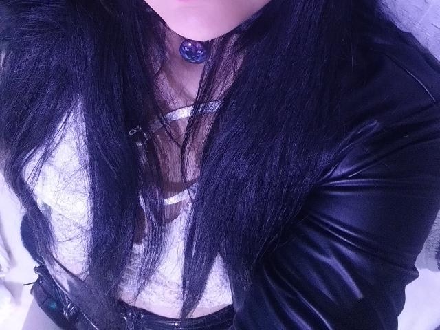 LindaSophieX with long black hair, wearing a white lace top and black leather jacket, purple gem necklace, showing her tits and a hint of her ass.