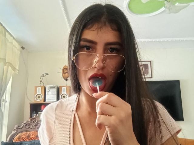 MadissonFontaine with long dark hair, wearing glasses, sucking on a blue lollipop, her red lips parted, in a pink cardigan, with a small nose piercing.
