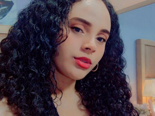 JackelineRoset with curly black hair, red lipstick, and hoop earrings, looking at the camera with a slight smile, soft blue background, and warm bedroom light.