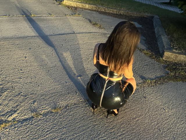 AnaHathor, crouching on a street, wearing a black latex skirt and boots, with a yellow rope tied around her waist, and long brown hair, in sunlight.
