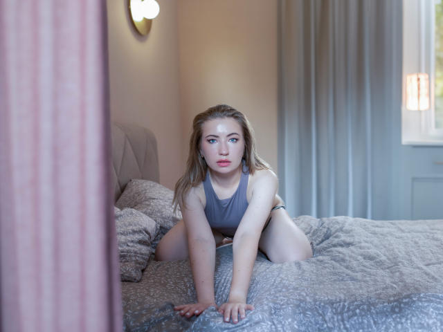 MiaFlirt est à quatre pattes sur un lit, vêtue d'un débardeur gris et d'une culotte noire, ses seins étant légèrement visibles, dans une chambre à coucher éclairée doucement..