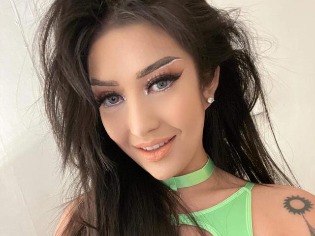 Yukie69 has long, messy dark hair, green choker, and a sun tattoo on her right shoulder. She's smiling with winged eyeliner, peach lips, and diamond earrings.