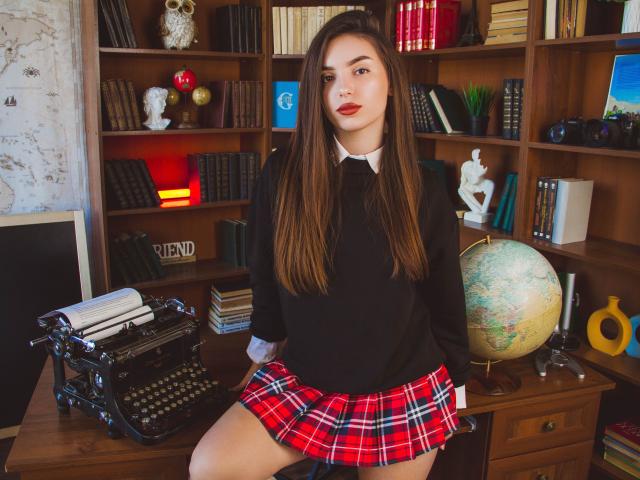 EmmaBettis, with long brown hair, red plaid skirt, black sweater, and white collar, sits on a wooden desk, showing her bare thigh and holding a globe.