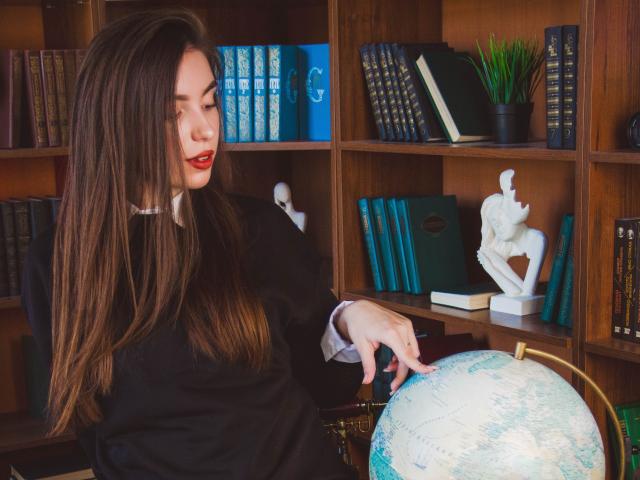 EmmaBettis with long brown hair, red lipstick, black dress, pointing at a globe in a wooden bookshelf, featuring books, a white statue, and a green potted plant.