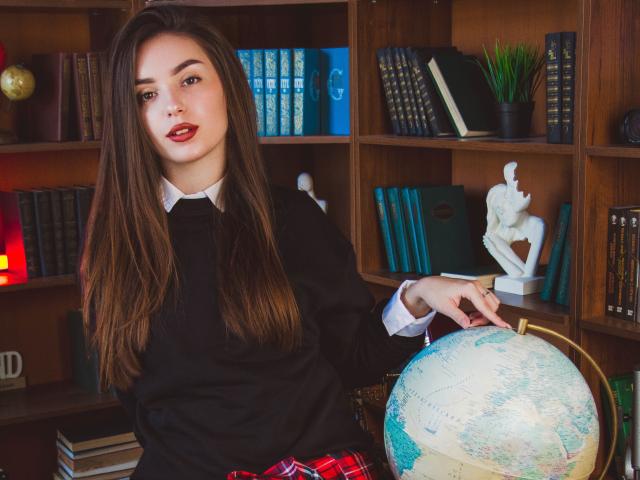 EmmaBettis with long brown hair, red lipstick, black sweater, white shirt, plaid skirt, and ring, holding globe in front of wooden bookshelf with statues and books.