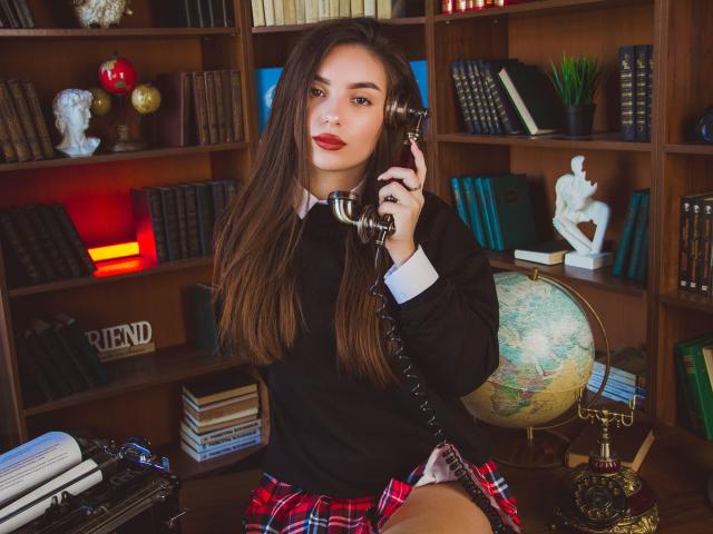 EmmaBettis with long brown hair, red lipstick, black sweater, and red plaid skirt, holding a vintage phone, sitting in a library, globe and white statue in background.
