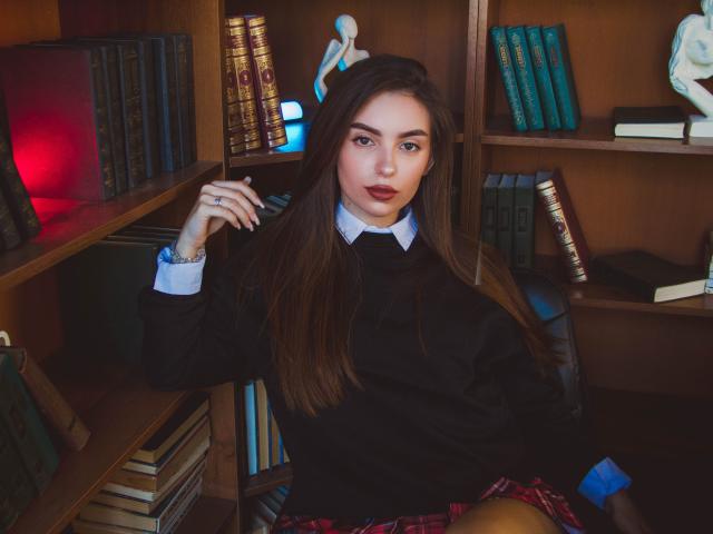 EmmaBettis with long brown hair, wearing a black sweater, white shirt, and red plaid skirt, sitting in a wooden bookshelf, looking at the camera with a slightly open mouth.