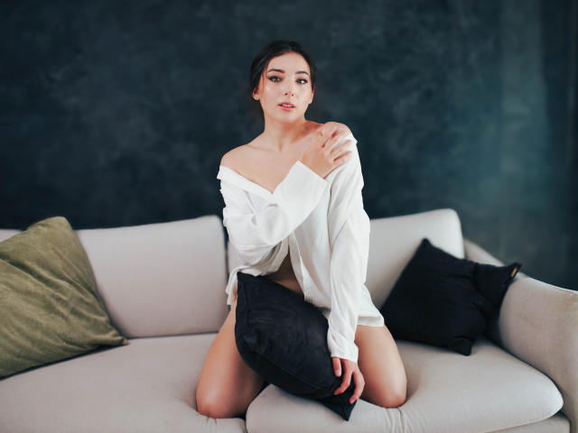 KissMegan with bare shoulders, white off-shoulder shirt, black pillow, and green pillow on white couch, kneeling, looking at camera. She has medium tits, dark hair, and fair skin.