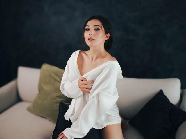 KissMegan with dark hair, wearing an off-shoulder white shirt, exposing her tits, sitting on a couch with green and black pillows, looking at the camera.