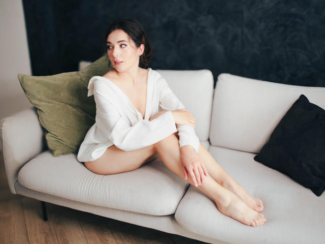 KissMegan, avec sa peau pâle, ses cheveux foncés et ses petites seins, est assise sur un canapé blanc, vêtue d'une chemise blanche ample, les jambes croisées, avec des coussins verts et noirs derrière elle..