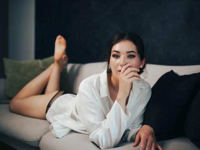 KissMegan, with dark hair and makeup, lies on a couch, wearing an open white shirt and black underwear, biting her finger, with her legs up, showing her pussy.