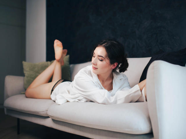KissMegan, avec ses cheveux foncés, allongée sur un canapé gris, portant une chemise blanche ouverte et des culottes noires, les jambes relevées, souriante, pieds nus, avec un mur noir en arrière-plan..