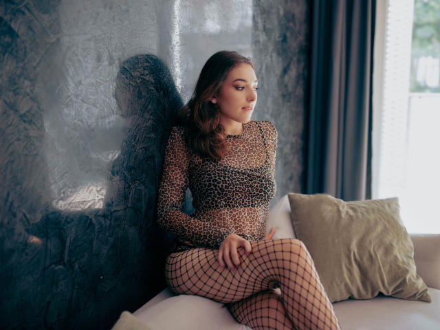 KissMegan with brown hair, wearing a leopard print dress and fishnet stockings, sitting on a white couch, looking to the side, her ass and tits visible.