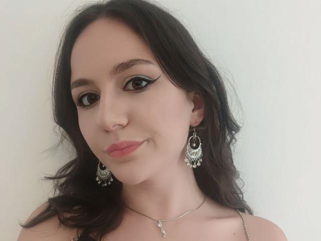 AshleyCrystal, com cabelos escuros e ondulados, usando brincos e colar de prata, tem delineador nos olhos, lábios rosa e um leve sorriso. Seios não visíveis, foco no rosto e parte superior do peito..