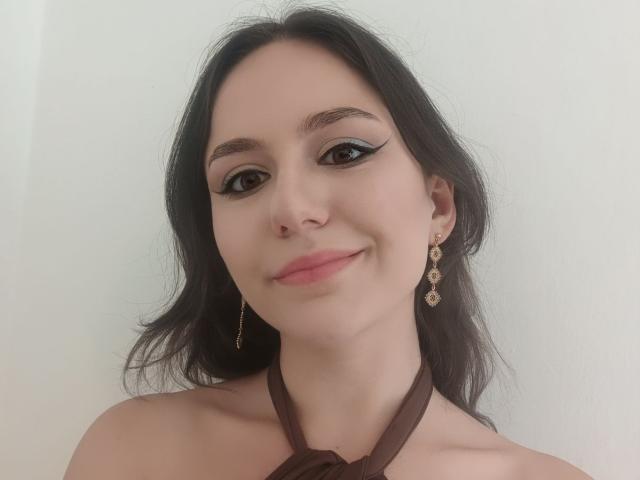 AshleyCrystal hat dunkles Haar, schwarzen Eyeliner, rosafarbene Lippen und ein leichtes Lächeln. Sie trägt goldene Ohrringe und ein braunes Halstuch-Top. Ihre Brüste sind teilweise sichtbar..