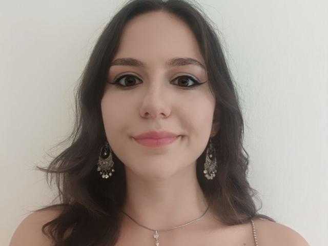 AshleyCrystal hat dunkles, welliges Haar, braune Augen mit dicker schwarzer Eyeliner, rosafarbenen Lippenstift und trägt kunstvolle silberne Ohrringe mit einer kleinen Anhänger-Kette und lächelt sanft..