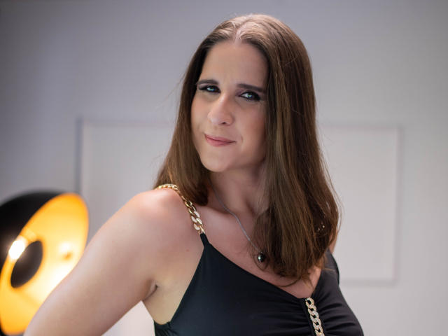 KayaBeauty has medium brown hair, black sleeveless top with gold chain, subtle smile, and blue eyes, standing with her arm slightly raised, showing her tits.