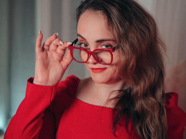 AdelaideRosabel with red glasses, red dress, and wavy brown hair, holding glasses with her right hand, smiling slightly, red lipstick, and white nails.