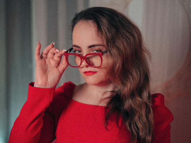 AdelaideRosabel with wavy brown hair, red lipstick, and red glasses, holding them with her right hand, wearing a red off-shoulder top, looking slightly to the side.