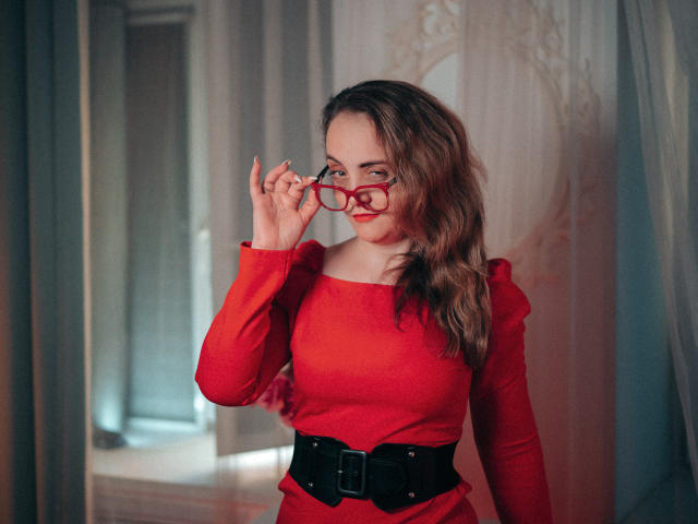 AdelaideRosabel with wavy brown hair, wearing a red long-sleeve dress and black belt, holding red glasses and smiling. Her tits are slightly visible through the dress.