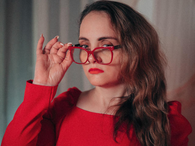 AdelaideRosabel with wavy brown hair, red lipstick, and red glasses, holding them with her right hand, wearing a red long-sleeve top, looking at the camera.