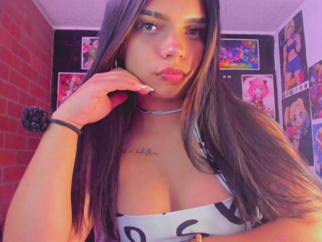 EmilyCandyNaughty with long brown hair, pink lipstick, and a silver choker, holding her cheek, showing her tits in a white dress with black letters, in a pink-lit room.
