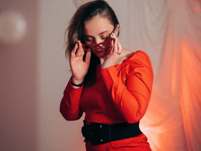 AdelaideRosabel with red dress, black belt, and red sunglasses, adjusting them with hands near face, looking down, wearing a ring on her right hand, in a soft, warm light.
