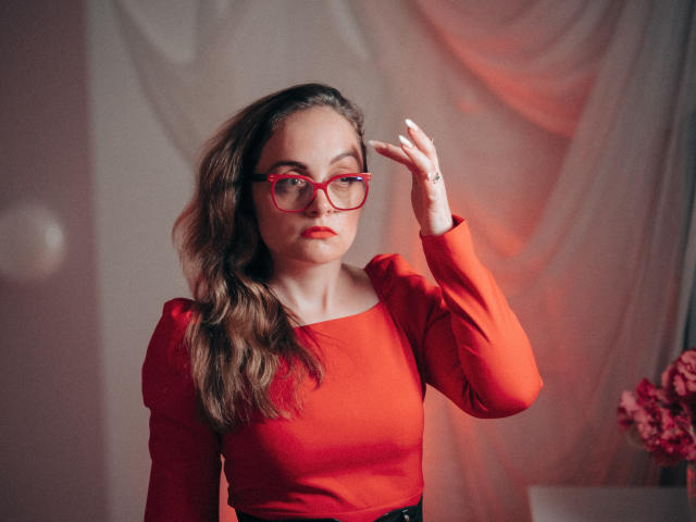 AdelaideRosabel with wavy brown hair, red glasses, and red long-sleeve top, adjusting her glasses, has red lipstick and a ring on her right hand.