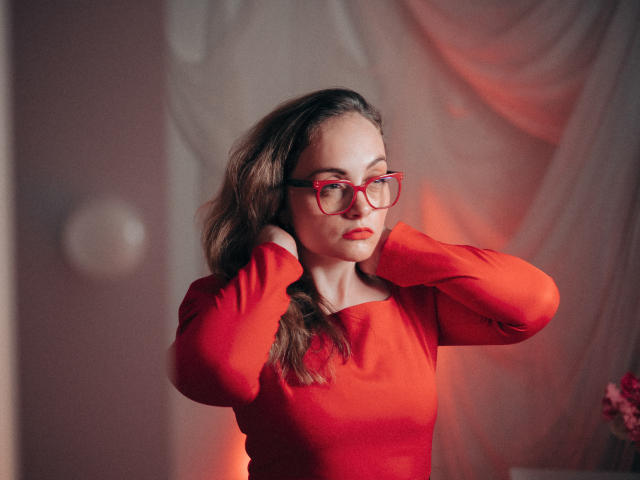 AdelaideRosabel with red glasses, red long-sleeve top, and brown wavy hair, hands in hair, eyes closed, red lipstick, soft lighting, red and pink hues.