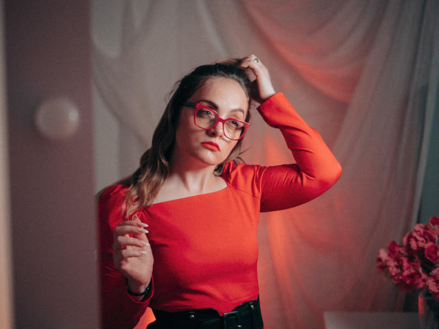 AdelaideRosabel with red glasses and red long-sleeve top, hand in hair, looking at camera, standing, pinkish-red light, black belt, flowers in background, soft lighting, relaxed pose.