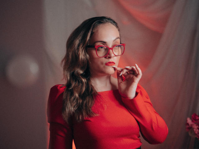 AdelaideRosabel with wavy brown hair, red glasses, and red dress, biting her finger, looking at camera, red lips, soft light, background blurred, red and pink hues.