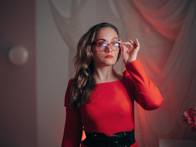 AdelaideRosabel with wavy brown hair, red glasses, and red long-sleeve top, adjusting glasses, wearing black belt, red lips, and subtle makeup. Soft lighting.