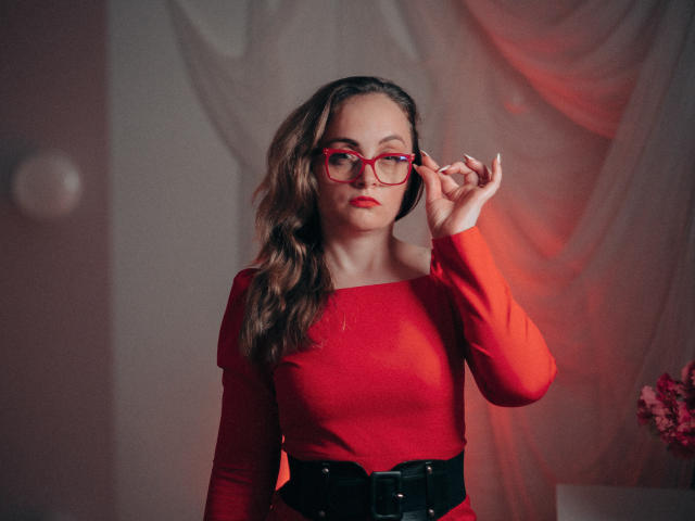 AdelaideRosabel with wavy brown hair, red glasses, red long-sleeve dress, black belt, adjusting glasses, red lipstick, soft light, slight blush, confident expression, medium tits, pretty face.