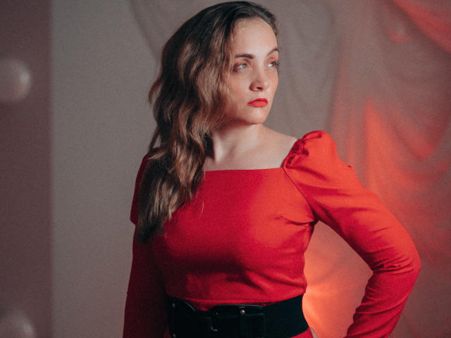 AdelaideRosabel with wavy brown hair, red dress, and black belt, looking to the side with red lipstick, showing her tits subtly. Red and warm lighting.