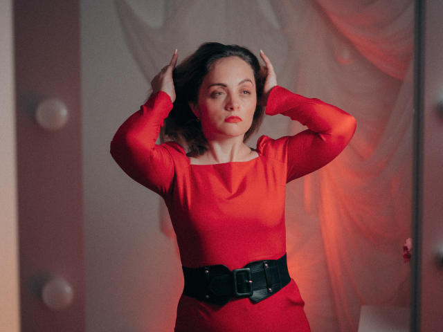 AdelaideRosabel with hands in her brown wavy hair, wearing a red long-sleeve dress and black belt, standing with a serious expression in a softly lit room.