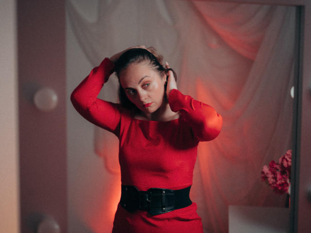AdelaideRosabel in a red dress with black belt, hands in hair, red lipstick, looking at camera, medium shot, soft light, slightly blurred background, serious expression, red and pink hues.