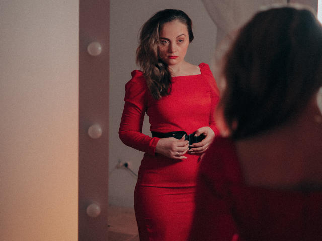 AdelaideRosabel with wavy brown hair, wearing a tight red dress, standing in front of a mirror, holding black camera, looking serious, her tits slightly visible through dress.