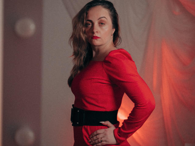 AdelaideRosabel with wavy brown hair, red dress, black belt, hands on hips, red lipstick, serious expression, standing in dimly lit room, soft red light in background.