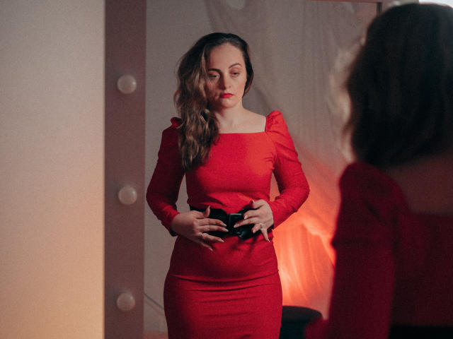 AdelaideRosabel in a red dress, standing, hands on black belt, looking at reflection, wavy brown hair, red lipstick, soft lighting, mirror, warm red and pink tones.