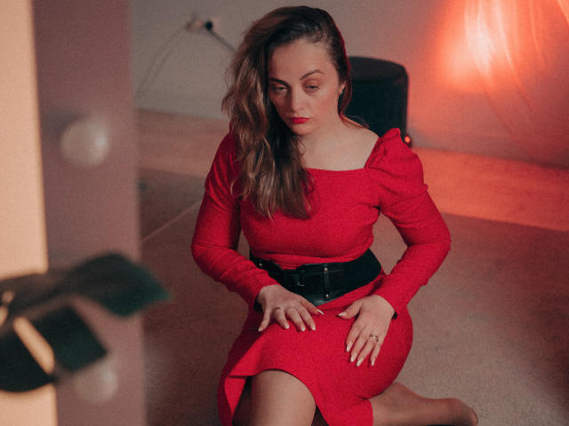 AdelaideRosabel with wavy brown hair, red dress, black belt, and red lipstick, kneeling, looking down, hands on her thigh, soft lighting, subtle shadow.