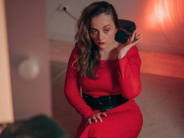 AdelaideRosabel with wavy brown hair, red dress, and black belt, sitting, hand near face, looking at camera, red lighting, soft shadow, sexy, confident, subtle smile.