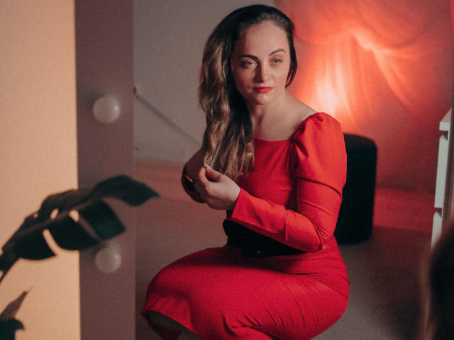 AdelaideRosabel in a red dress, sitting with wavy brown hair, holding a small black purse, smiling slightly, with a red light in the background. Boobs, ass, pussy not visible.