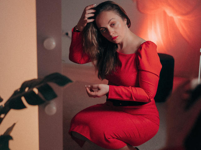 AdelaideRosabel in a red dress, sitting with one hand in her brown hair, looking at the camera with a slight frown, her ass and tits partially visible.