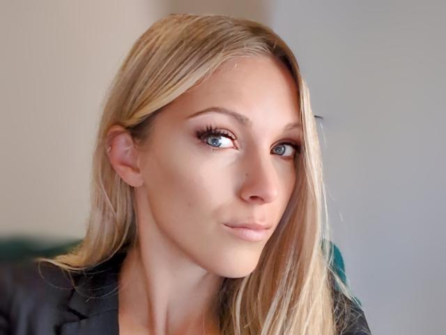 HelenaHot69 has blonde hair, blue eyes, and a slight smirk. She's wearing a black leather jacket, with her face and upper shoulders in focus.