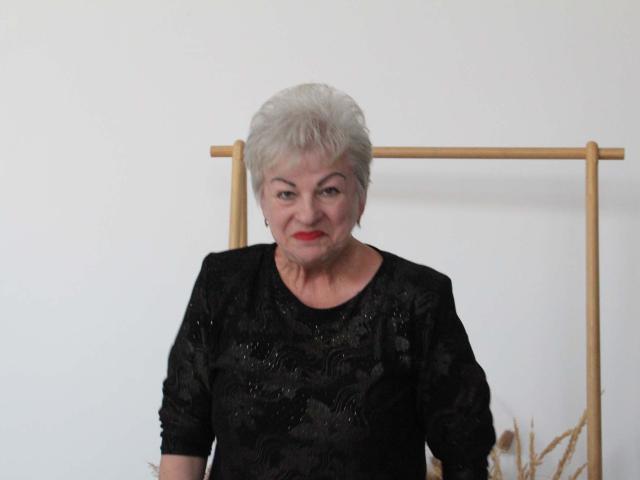 KayleSmoll has short white hair, red lipstick, and wears a black lace top, standing in front of a wooden frame. Her tits are small, and she looks confident.