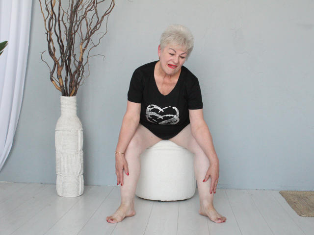 KayleSmoll, short white-haired woman, sitting on white ottoman, black t-shirt with white snake print, bare legs, red nails, white vase with brown branches, grey wall, white floor.