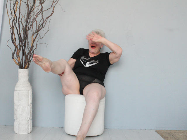 KayleSmoll, elderly woman with white hair, black t-shirt, and black panties, sitting on white cylinder, leg raised, covering face, bare feet, white vase with dry branches on left.